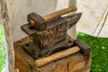 Traditional anvil with wooden hammer, set on a tree stump under a tent. Retro blacksmith tools used in crafts and metalworking. Photo taken outdoors, against a background of white canvas and green gra