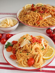 Plate of spaghettini with cherry tomato with serving bowl of the same, bowl of shaved parmesan and fresh cherry tomatoes on-stem in the background