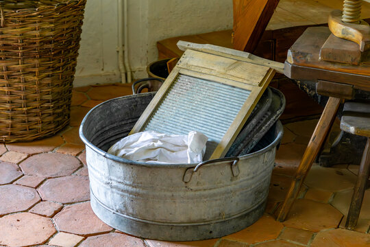 Close-up of an old-fashioned galvanized tub with a washboard and white fabric in the interior of a rustic room. In the background, a wicker basket and wooden furniture create a vintage atmosphere.