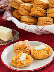 Two sweet potato biscuits, one open with butter, in front of a basket with a group of them and a stick of butter in a dish in the backgroun