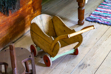 wooden, antique baby carriage placed on a wooden floor in an interior stylized as a traditional cottage. Next to it is a fragment of a wooden stool and a colorful, braided rug.
