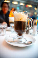 Close-up of a freshly served Viennese coffee with whipped cream in a clear glass mug, set on a white plate at a stylish European café. Perfect for food, travel, and holiday lifestyle visuals