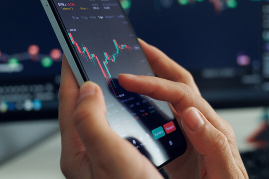 A person's hands operate a smartphone displaying a dynamic stock market chart with fluctuating red and green bars, against 