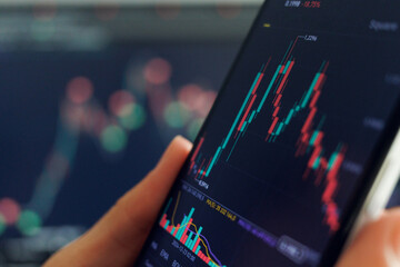 A hand holds a smartphone showing a fluctuating stock chart with red and green candlesticks. Another blurred screen displays similar data in the background.