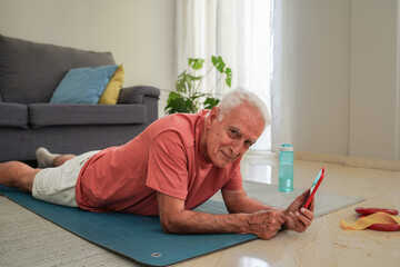 Elderly man using a smartphone while relaxing on a yoga mat and smiling at camera at home