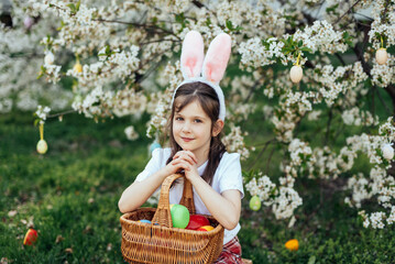 Easter egg hunt. Girl child Wearing Bunny Ears Running To Pick Up Egg In Garden. Easter tradition. Baby with basket full of colorful eggs.