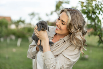 A happy woman smiles while holding a fluffy rabbit in a green backyard, embodying joy and affection for pets.