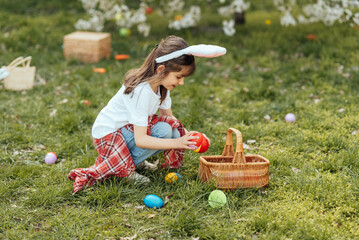 Easter egg hunt. Girl child Wearing Bunny Ears Running To Pick Up Egg In Garden. Easter tradition. Baby with basket full of colorful eggs.