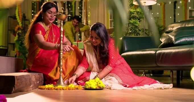 Decorating flower rangoli around samai, Indian young girl senior mother in traditional wear joyfully preparing for Hindu festival Diwali celebration indoors in evening with diya and colorful design