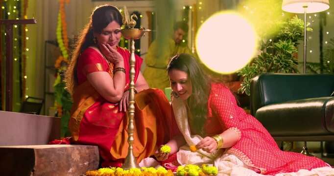 Decorating flower rangoli around samai, Indian young girl senior mother in traditional wear joyfully preparing for Hindu festival Diwali celebration indoors in evening with diya and colorful design