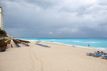 Praia de Cancún com cadeiras e guarda-sóis sob céu escuro e nublado, mar azul-turquesa antes de uma tempestade.