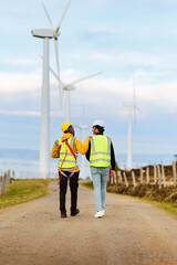 Obraz premium Two engineers in safety gear walk along a path in a wind turbine park, discussing renewable energy solutions amid towering turbines