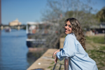 Young, pretty, blonde woman, wearing shirt, posing looking at camera, leaning on railing with river...