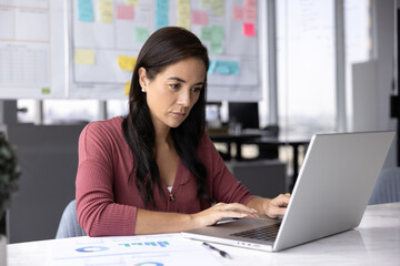 Data review and analysis. Busy millennial female office worker financial analyst accountant sit at workplace focused on preparing report on notebook engaged in digital paperwork using corporate app