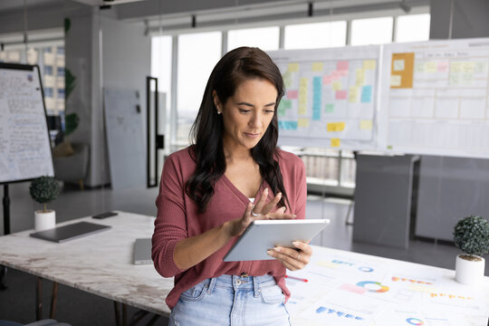 Modern tech as creative environment tool. Young businesswoman use digital tablet in office boardroom with flipchart agile board statistic data on table managing project online review digital documents