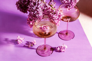 Two crystal glasses with sparkling wine and lilac flowers on violet background, decorated with scattered blossoms