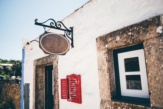 Close-up of a rustic blank hanging shop sign mockup on a traditional whitewashed building with stone framing, red shuttered menu window, and vintage lamp under clear blue sky