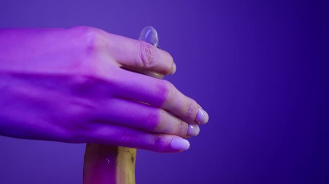 Woman's hands gently and gently place a rubber condom on a yellow phallic banana, stretching it along its entire length in a neon erotic purple color