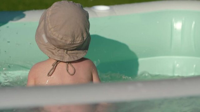 Baby boy stands with his back, splashing in backyard, slow motion. Infant child in stylish sun panama hat standing backside in inflatable pool, embodying summer and creating unforgettable memories