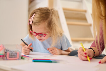 child and adult engaging in drawing activity together. young girl with pink glasses and hairclip focused on coloring with crayons, expressing creativity and concentration