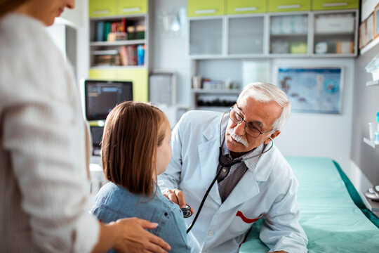 Smiling doctor using stethoscope to check young girl at medical clinic with mother nearby