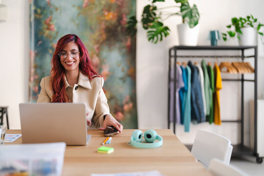 An Arab woman smiles while working on her laptop. She is in a bright, modern office space with plants and a clothing rack. - Powered by Adobe