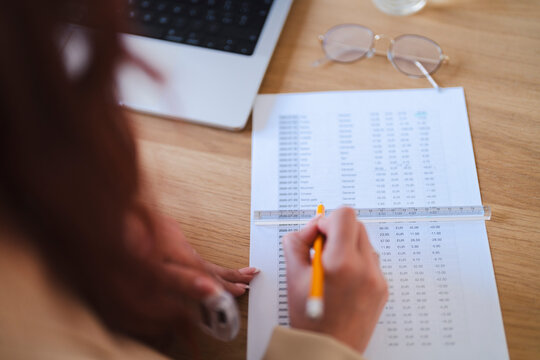 An Arab female reviews financial data. She uses a ruler and pencil to analyze a printed spreadsheet at her desk with a laptop and glasses nearby.