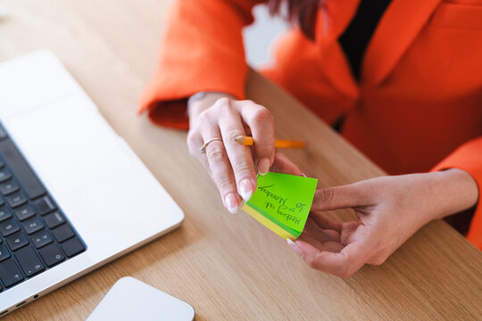 An Arab woman reviews her schedule. She is holding a sticky note with "Meeting at 10 Monday" written on it, sitting at her desk with a laptop.