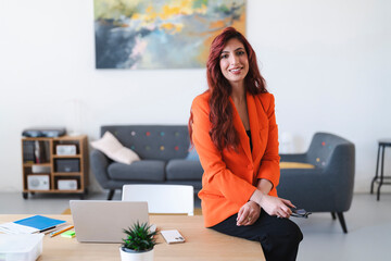 Confident Arab businesswoman in an orange blazer sits on her desk. She is holding glasses and smiling in her modern office space.