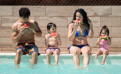 Front view of happy latin family eating slices of refreshing watermelon, sitting by the pool with...