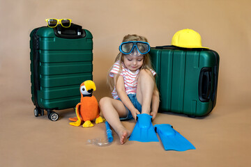 Funny little girl sitting in the middle of a green suitcase with a toy parrot with a diving mask on her head, putting on flippers, dreaming of a trip to the sea. Children's vacation, travel, flight. 