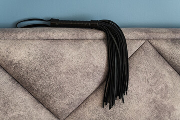 A black leather flogger for BDSM practices lies on a soft, textured grey headboard in front of a blue wall.