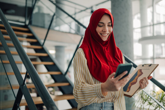Smiling businesswoman wearing hijab using smartphone and holding clipboard in modern office