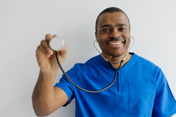 Smiling African American doctor in blue scrubs holding a stethoscope close to the camera lens in playful gesture, happy young black male medical worker standing on white studio background, closeup