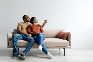 Split system. Cheerful young black couple opening air conditioner with remote while sitting on couch over white studio background, happy african american man and woman using climate control
