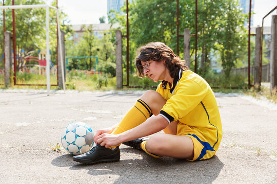 Teen boy in yellow soccer uniform sitting on asphalt and tying shoelaces before training