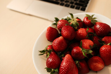 A white bowl brimming with ripe red strawberries sits beside an open silver laptop on a light wood desk, blending vibrant fruit with modern tech in a clean, inviting workspace scene.
