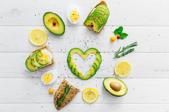 Avocado slices arranged in a heart shape surrounded by assorted produce including Avocados, hard boiled eggs, bread, fruit and herbs