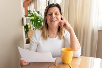 Mature woman working on documents, sipping coffee while seated at wooden dining table, relaxed home...