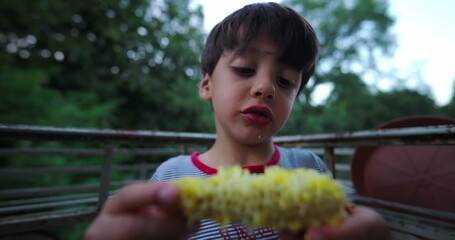 Close-up of a boy holding corn on the cob and examining it, focused on the corn with a calm outdoor backdrop during a summer evening