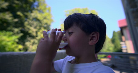 Obraz premium Young boy drinking milk outdoors with blurred trees in the background on a sunny day, close-up of the side profile. Focus on the child holding a glass of milk against a natural backdrop