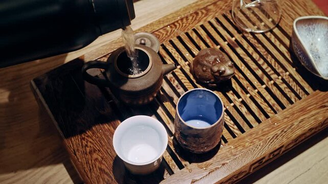 A tea master begins the brewing process by pouring boiling water from a thermos into a clay teapot filled with tea leaves, set on a wooden tray with cups.