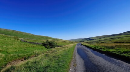 Fototapeta premium Scenic Country Road Cuts Through Lush Green Hills Under A Clear Blue Sky, Inspiring Travel And Adventure.