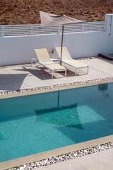 A swimming pool surrounded by a minimalistic patio features two beige sun loungers and a large umbrella