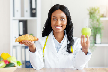 Making healthy food choices. Beautiful female nutritionist holding croissant and apple at table in...