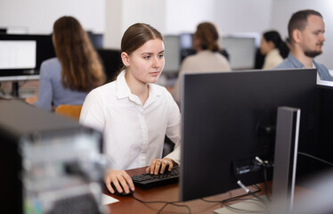 Female student of computer courses attentively looks at monitor screen, types on keyboard and...