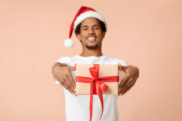 Celebrating winter holidays and gift from Santa Claus. Cheerful african american young man in hat gives box with present and looks at camera, isolated on light background, empty space, studio shot