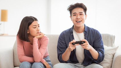 Gaming Addiction Problem. Discontented Asian Wife Looking At Infantile Husband Playing Video Game Sitting On Couch At Home. Computer Games, Waste Of Time, Male Emotional Immaturity Concept