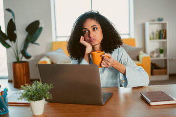 Young woman with curly hair looking thoughtful while holding coffee mug and sitting at desk with...