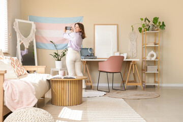Young woman taking picture of hanging transgender flag on beige wall at home, back view